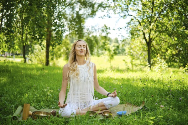 A woman sits cross-legged on a mat in a lush green park, eyes closed and arms resting on her knees.