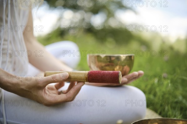 A person is holding a red-tipped mallet next to a singing bowl, seated on grass in a serene outdoor