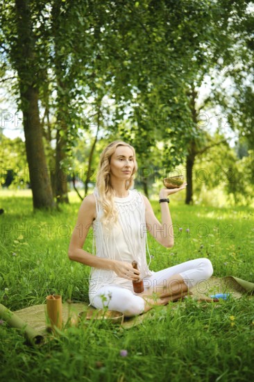 A woman practices meditation outdoors, holding a singing bowl while seated on a mat in a field. The