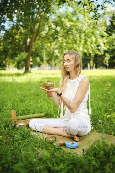A woman sits cross-legged in a grassy park, holding a singing bowl while meditating. Surrounded by