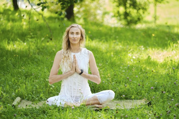 A woman dressed in white sits cross-legged on a mat, practicing yoga outdoors. Surrounded by