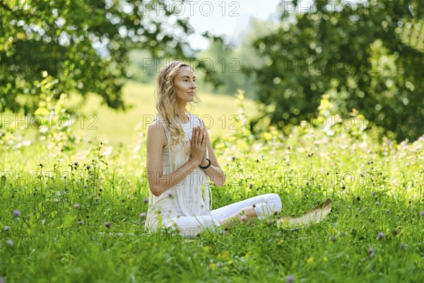 A woman sits cross-legged on soft grass in a vibrant green field. She closes her eyes and brings