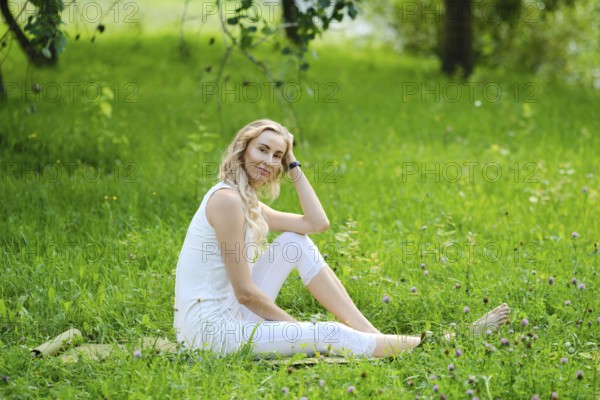 A young woman sits comfortably on a blanket in a lush green park. She has wavy hair and wears a