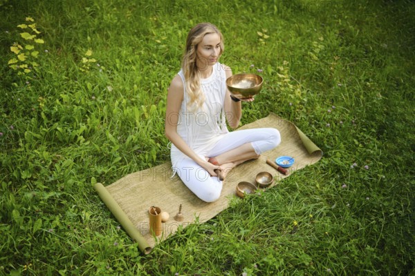 A woman sits cross-legged on a mat in a lush green field, holding a singing bowl. She is fully
