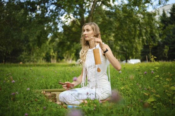 A woman sits cross-legged on the grass, surrounded by greenery, gently striking singing bowls with