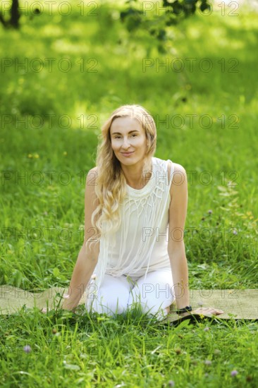 A woman with long hair sits on a yoga mat in a vibrant green park. She wears a light outfit and