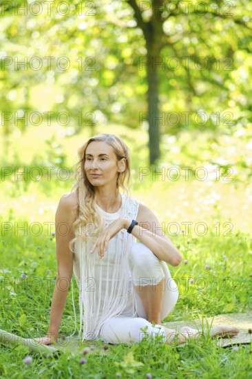 A woman with long hair sits elegantly in a vibrant green park. Dressed in white, she poses