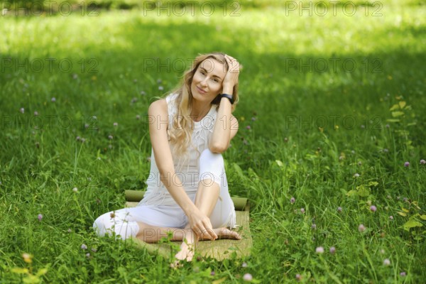 A young woman dressed in white relaxes on a mat in a lush green field. She smiles gently while