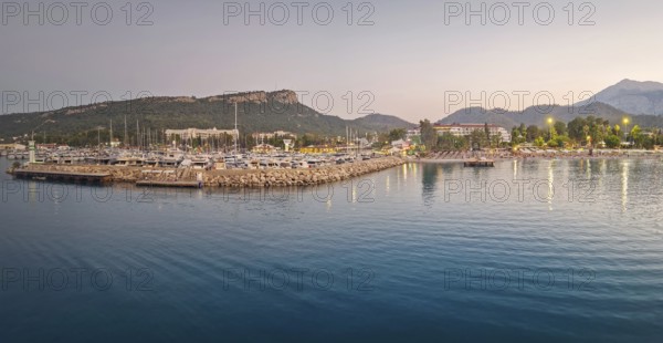 Kemer marina at dusk, filled with multiple docked boats. Lights from the harbor and hotels on the