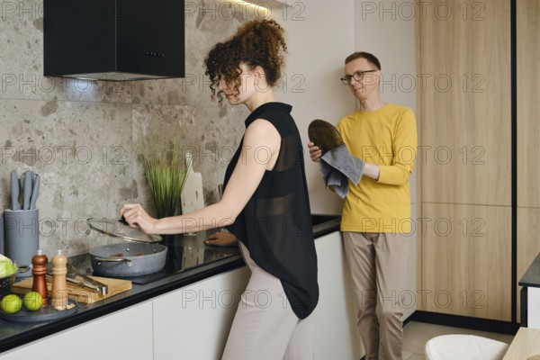 A woman stirs a pot on the stove while a a man wipes the washed dishes and watches her in a stylish