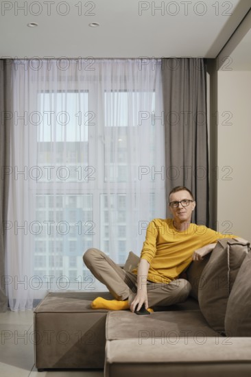 A man wearing a yellow shirt and socks and beige pants sits comfortably on a brown couch, relaxed
