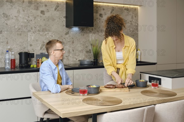 A woman slices roasted duck breast on a cutting board while a man sits at the dining table. They