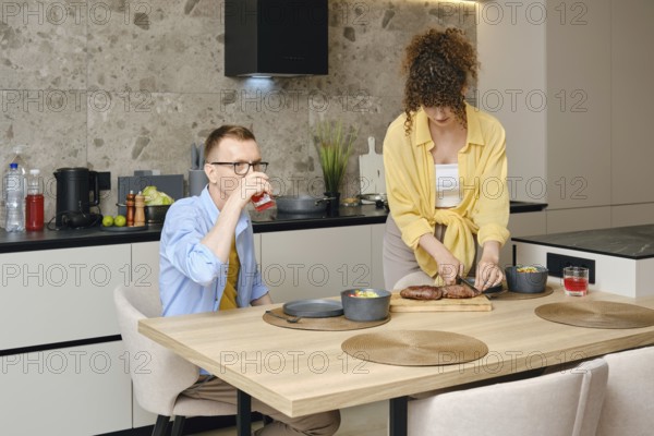 A man sits at a wooden dining table, sipping a drink, while a woman prepares a meal on the