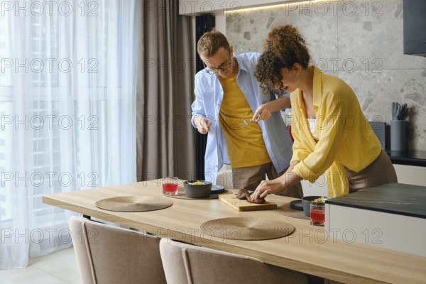 In a bright kitchen, a couple is joyfully preparing a meal together. The woman slices meat on a