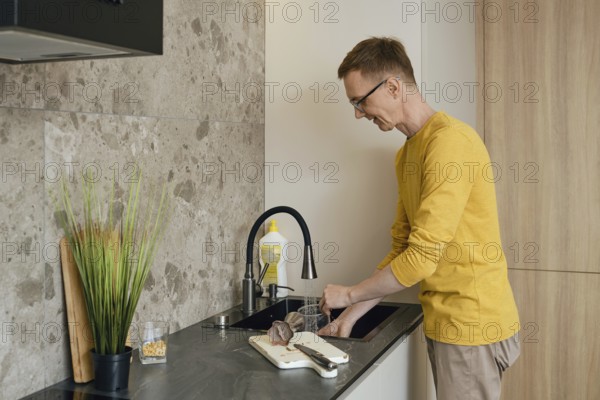 A man in a yellow long-sleeve shirt is washing utensils at a sleek kitchen sink. Natural light