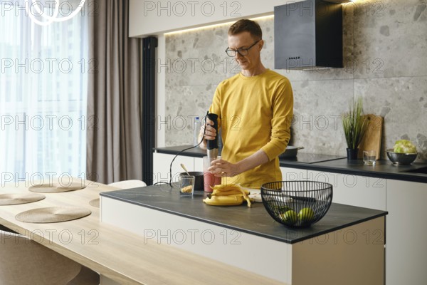 A middle-aged man prepares banana and strawberry smoothies in the kitchen using an immersion