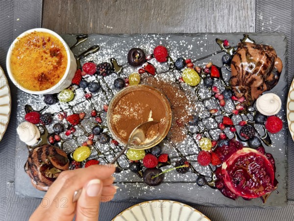 Hand with spoon tasting a dessert, surrounded by artfully arranged berries and chocolate Lower