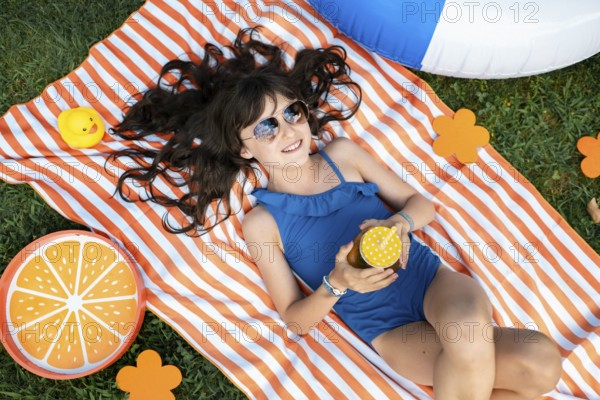 Girl, 10, with sunglasses in a blue swimming costume, lying relaxed in the meadow with a cocktail