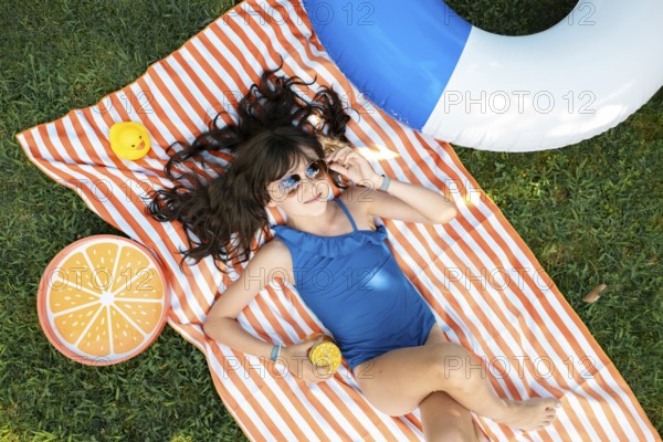 Girl, 10, with sunglasses in a blue swimming costume, lying relaxed in the meadow with a cocktail