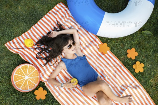 Girl, 10, with sunglasses in a blue swimming costume, lying relaxed in the meadow with a cocktail