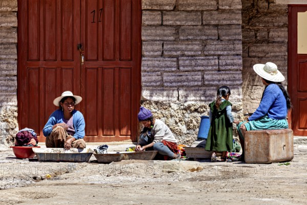 Women and children sit in front of a stone house, busy with housework. The sun shines brightly,
