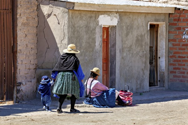 Woman and child in traditional dress standing in front of a brick house on a sunlit street,