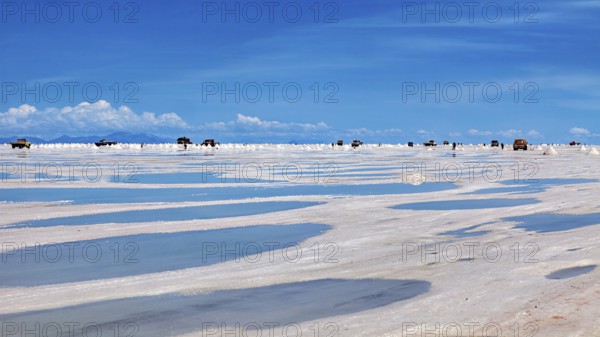 Puddles on a salt flat with vehicles on the horizon and clouds in the sky, The endless expanses of
