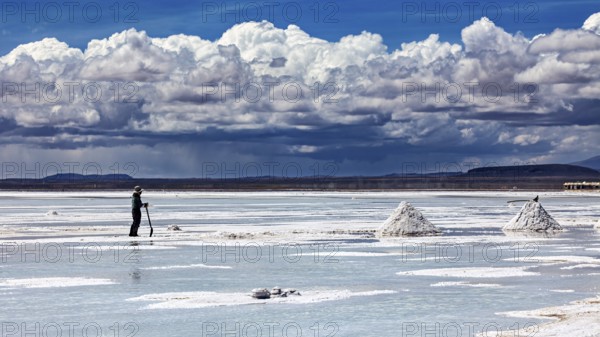 A single figure shovels salt under a dramatic cloudy sky on vast salt flats, The endless expanses