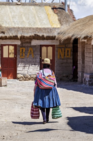 A woman in colourful traditional dress carries bags in a village with rustic buildings, Traditional