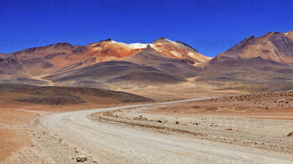 A winding sandy road through a dry landscape with reddish mountain peaks and blue skies, The