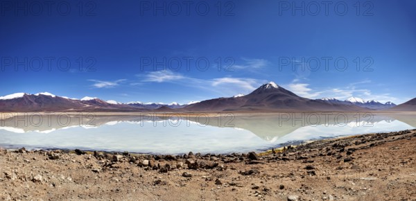 Wide view of a volcano with a reflecting lake and rocky shore under a blue sky, The Laguna Colorada