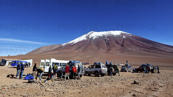 Groups of people and vehicles in a desert landscape with a snow-covered mountain under a blue sky,