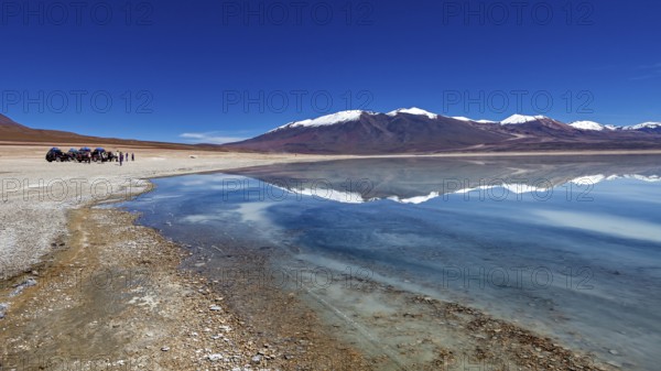 Panoramic view of a lake with snow-capped peaks, people and vehicles on the shore, The Laguna