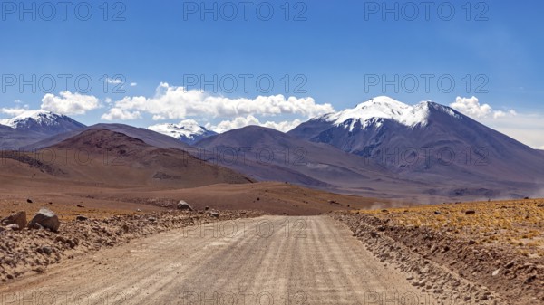 A straight, unpaved road leads through a barren mountain desert landscape on a sunny day, The