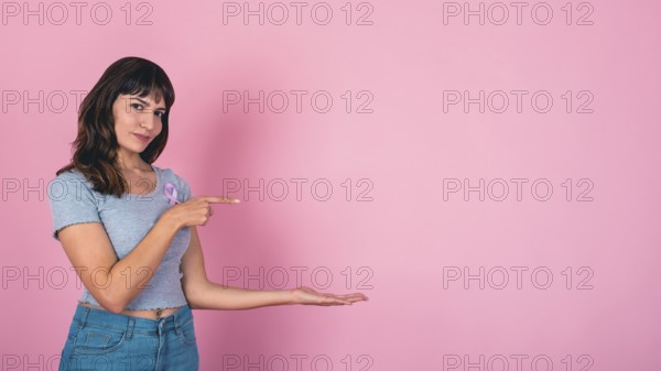 Young woman pointing and presenting with a pink ribbon for breast cancer awareness and copy space