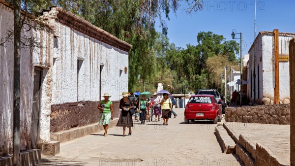 Pedestrians in summer clothes walk along a street with white buildings and a red car, The town of