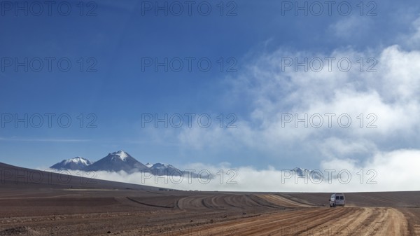 A dusty road leads through a remote desert landscape, bordered by mountains and clouds under a