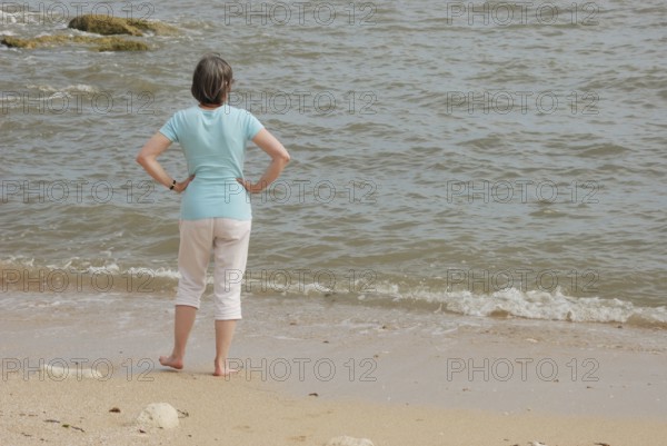 A woman stands on a sandy beach looking at the ocean waves, with rocks in the background in Royan