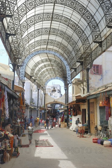 A vibrant market street with ornate metal canopy, bustling with people and colorful merchandise.