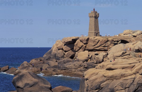 A lighthouse stands on a rocky coastline with people nearby and a clear blue sky. The granit rose