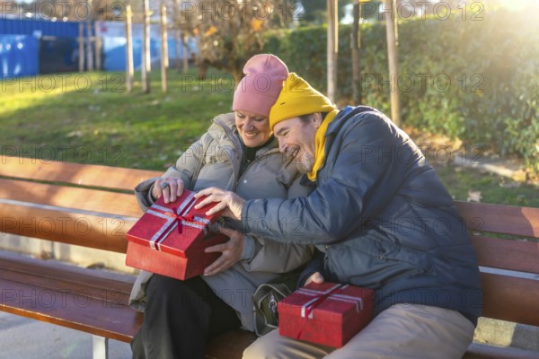 Elderly couple exchanging christmas gifts while sitting on a bench in a city park during a sunny