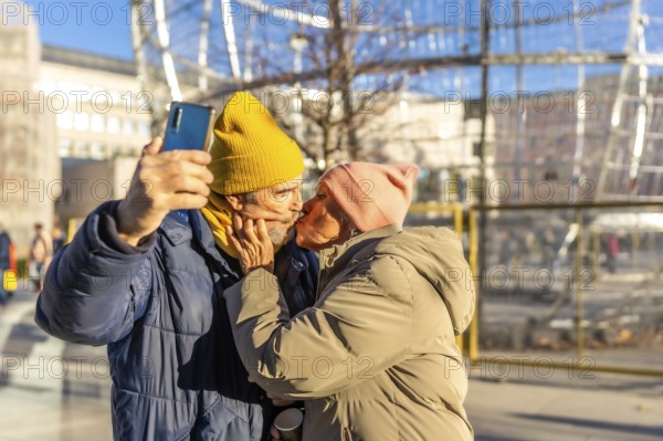 Happy senior couple kissing and taking a selfie with smartphone in a winter city during christmas