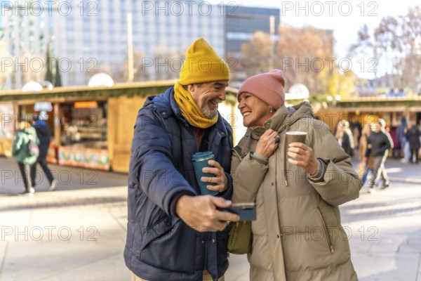 Smiling senior couple taking a selfie and drinking hot beverages at a christmas market in the city