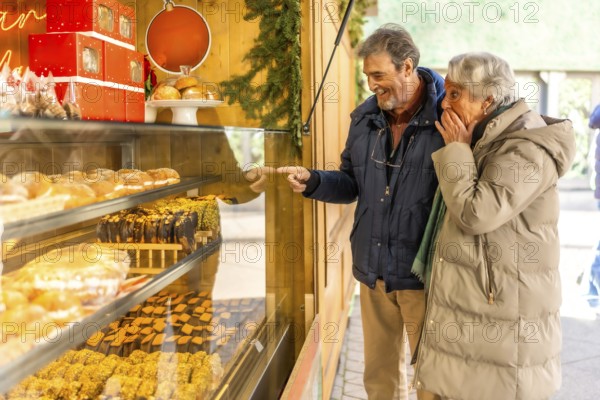 Joyful elderly couple pointing at delicious pastries in a bakery display window at an outdoor