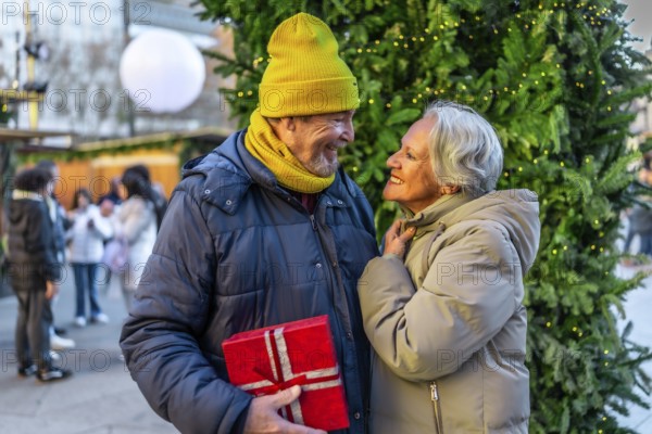 Elderly man surprising his wife with a christmas present at a christmas market during winter
