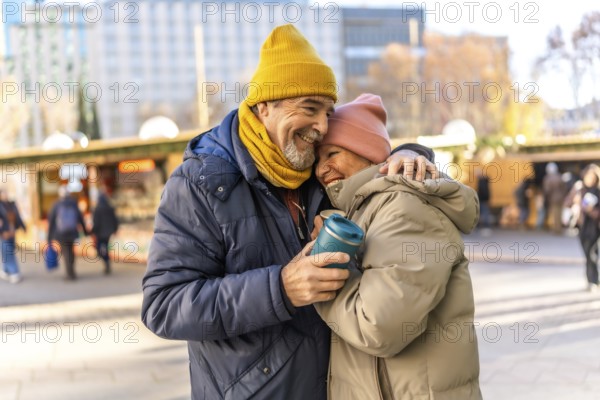 Elderly couple hugging and smiling at an outdoor christmas market, enjoying a warm beverage on a