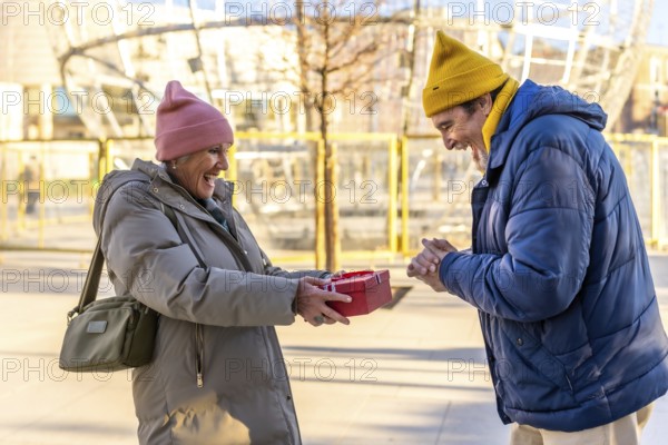 Cheerful senior woman giving a christmas present to her excited husband in a city during wintertime