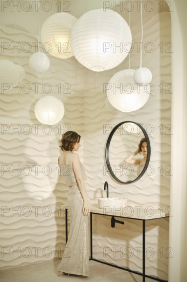 A woman dressed in a sparkling gown admires her reflection in a round mirror. Soft, textured walls
