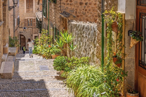 Alone woman walking in an idyllic cobblestone alley in an old village with green plants and flowers