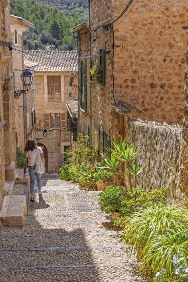 Woman walking in an idyllic cobblestone alley in an old village with green plants and flowers in